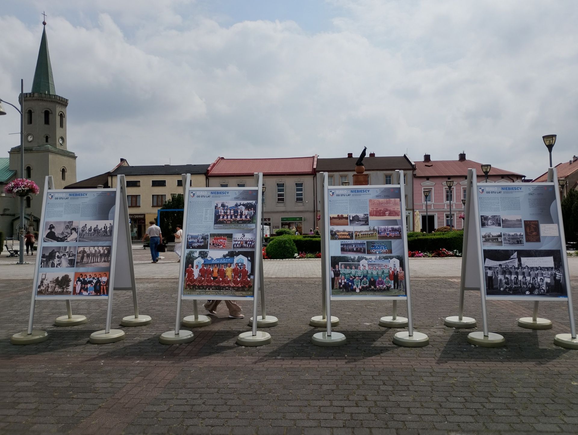 Zdjęcie plansz znajdujących się w plenerze. W tle kamienice rynku. Z lewej strony wieża kościoła.